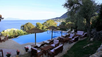 Zona de piscina exterior con sillones de descanso y sombrillas, con vistas a un cuerpo de agua y un paisaje montañoso.