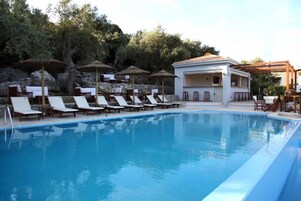 Una piscina con sillas de descanso y sombrillas, un edificio blanco con tejado de tejas, y árboles al fondo.