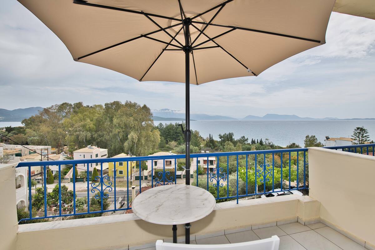 Un balcón con mesa y sombrilla, con vista a la costa, edificios y árboles.