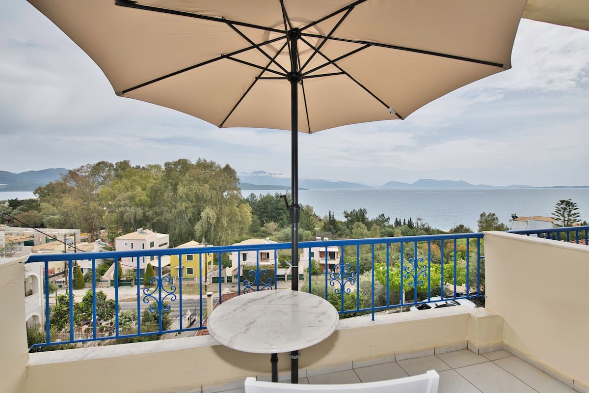 A balcony with a table and an umbrella, overlooking a coastal area with buildings and trees.