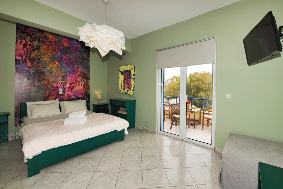 A bedroom with a large bed, a colorful abstract painting, a sliding glass door to a balcony, and a flat-screen TV mounted on the wall.