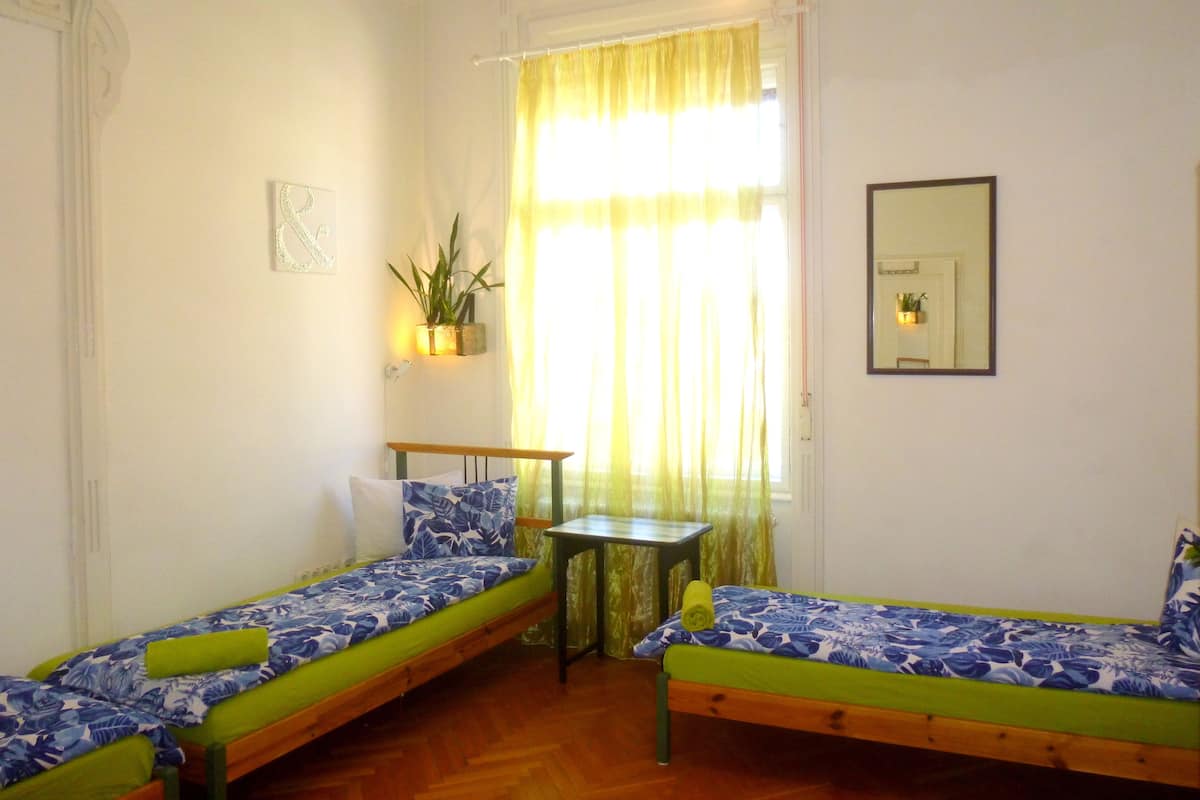 A small, bright room with two single beds, a small table, and a window with curtains.