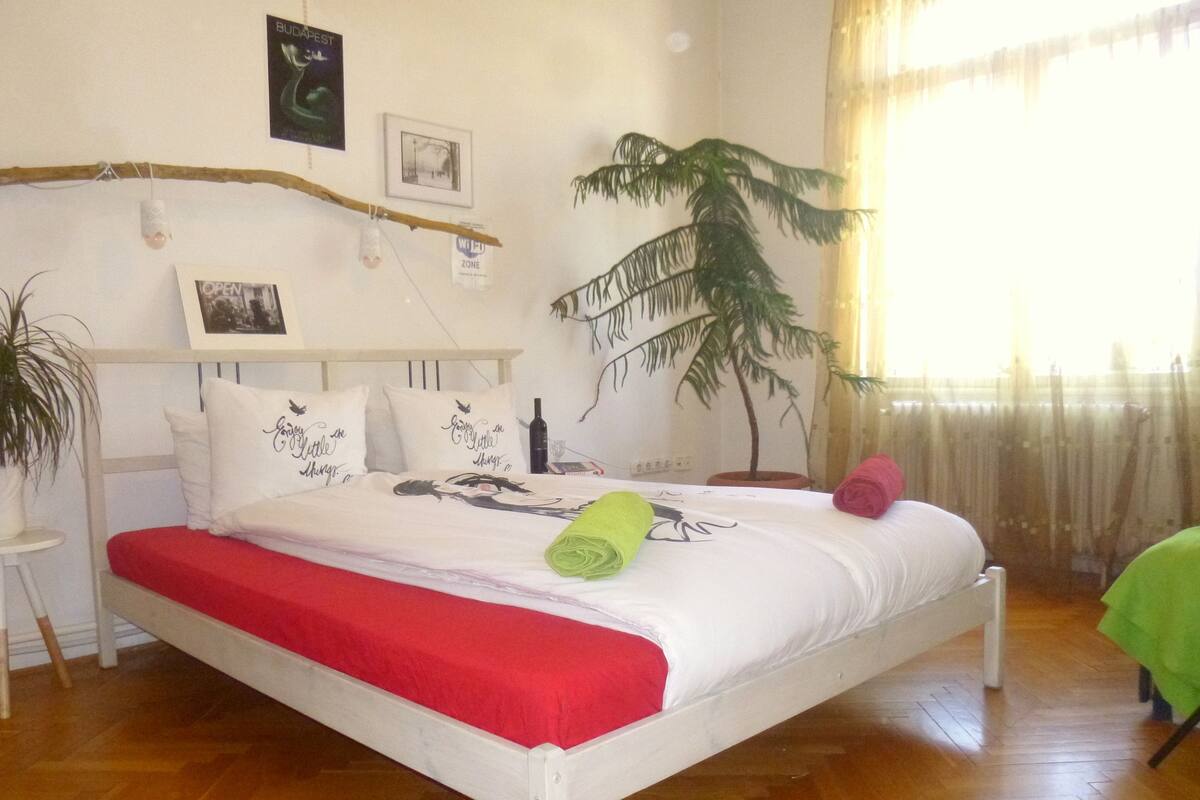 A bedroom with a white bed, red bedding, a small potted plant, a wall-mounted shelf, and a tropical plant decoration.