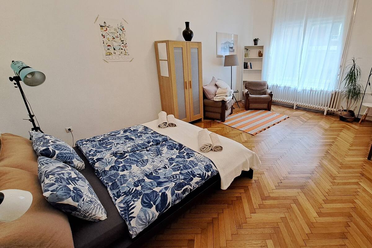 A bedroom with a wooden herringbone floor, a bed with a floral bedspread, a wooden wardrobe, a chair, a small table, and a radiator.