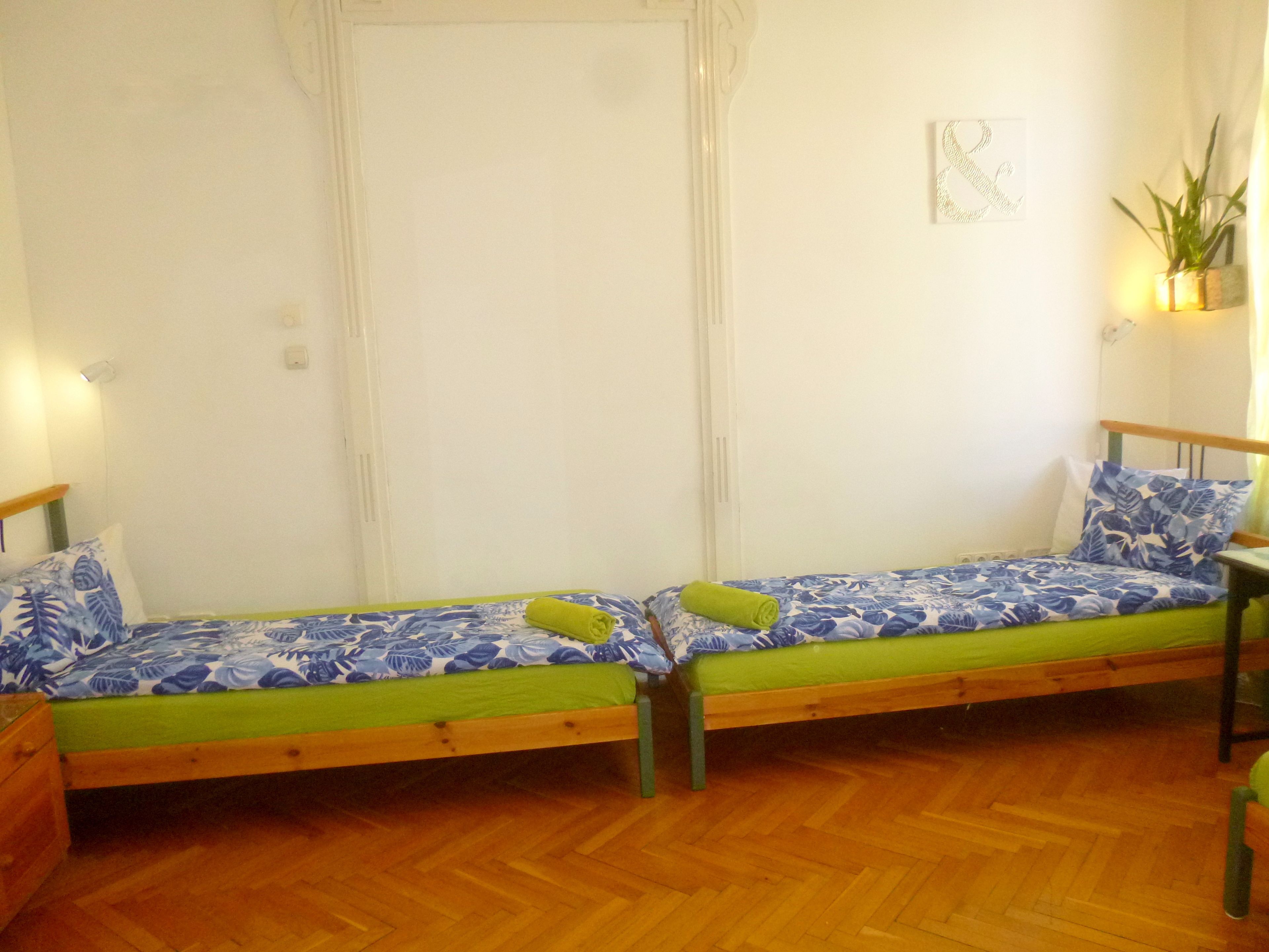 A room with two wooden beds with green and blue floral bedding, a wooden floor, and a wall-mounted light fixture.