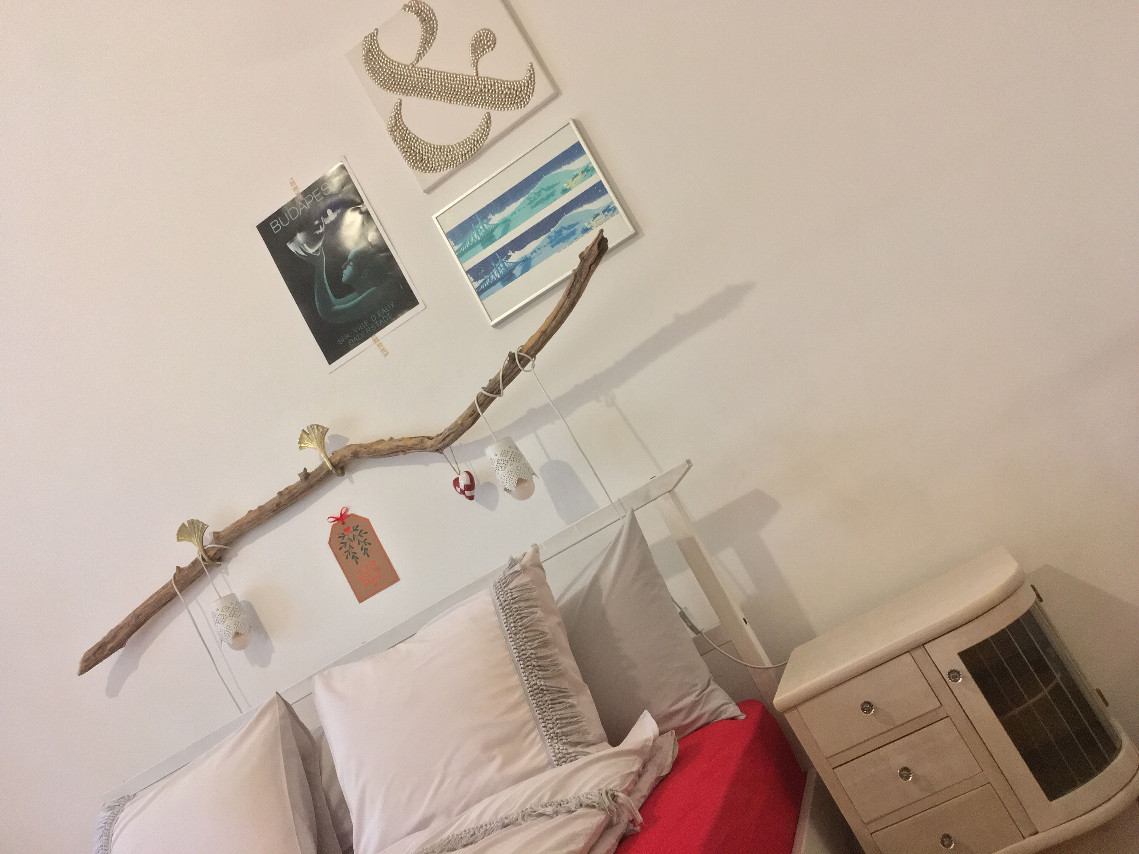 Quarto com cama, mesa de cabeceira, cômoda e decoração na parede, incluindo um galho, um colar e fotos emolduradas.