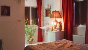 A bedroom with a bed, a bedside table with a lamp, a window with red curtains, and a potted plant. - Valentine Inn (Wadi Musa)