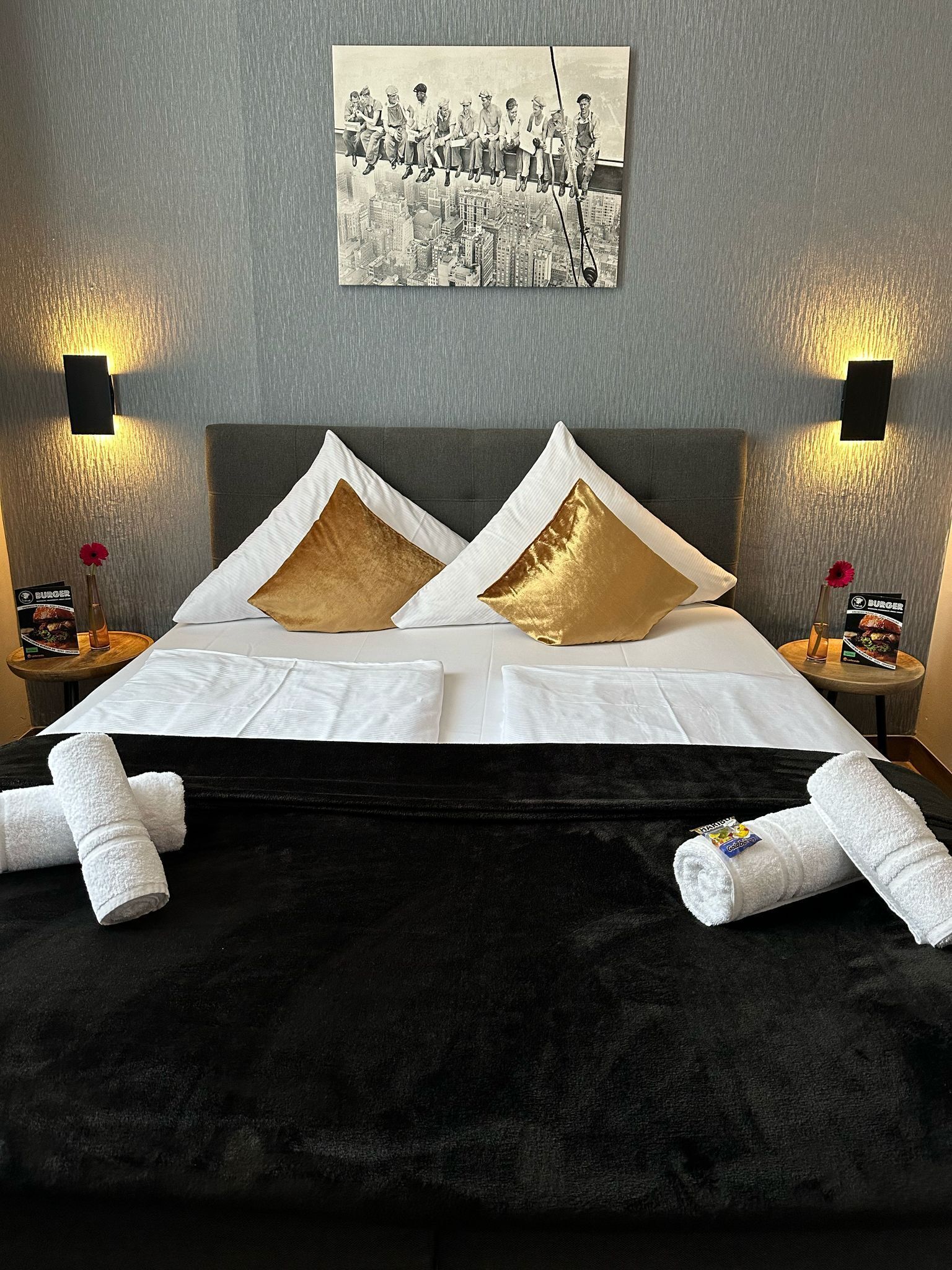 A hotel room with a large bed, two gold pillows, white linens, black velvet bedspread, two nightstands with lamps, and a black and white photo on the wall.