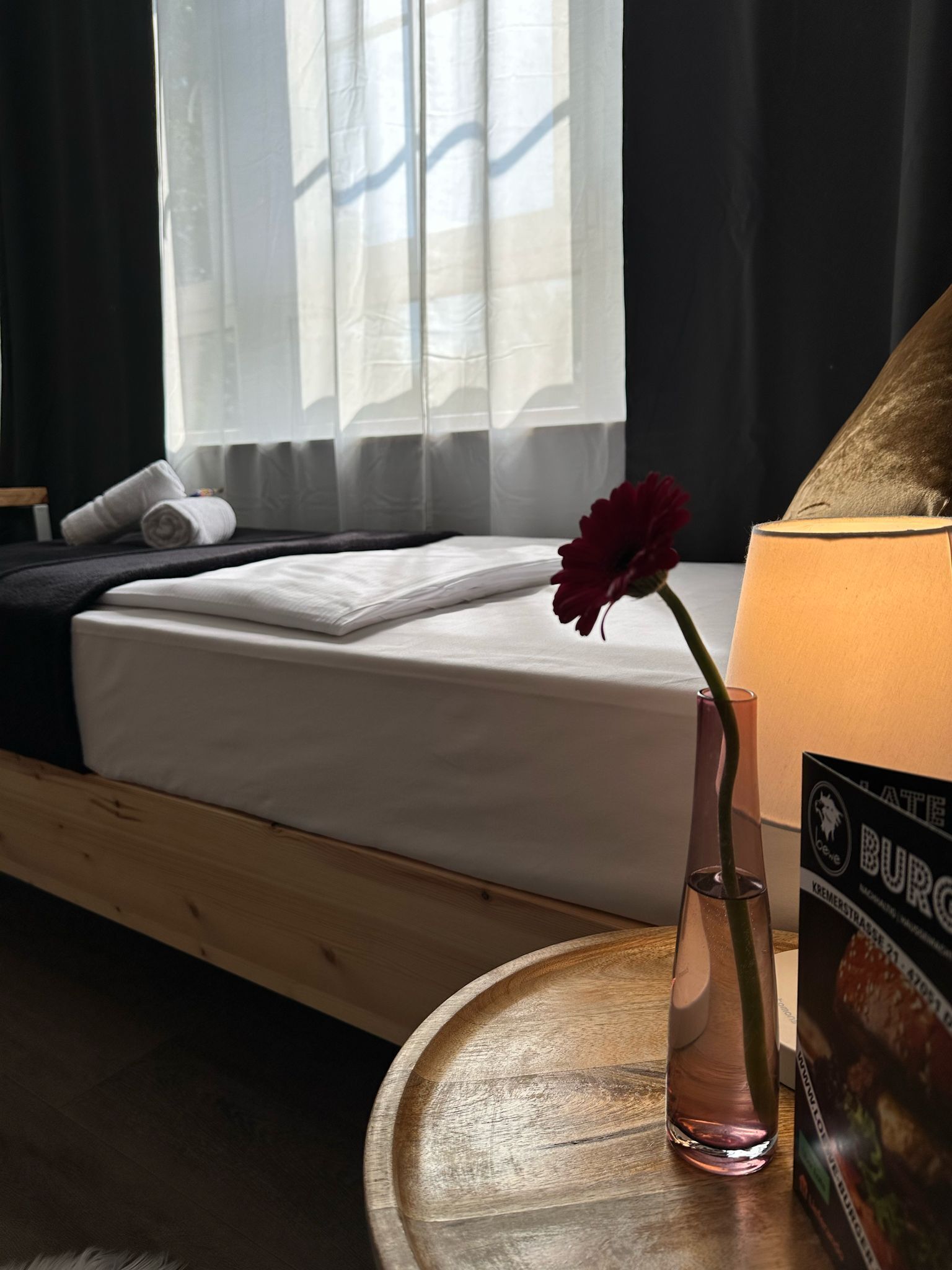 A hotel room with a bed, a vase with a flower, a table with a lamp, and a poster on the wall.