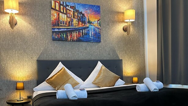 Una habitación de hotel moderna con una cama grande, dos lámparas de noche, un cuadro de un paisaje urbano y una cortina oscura.