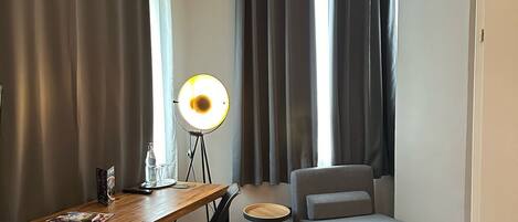 A hotel room with a bed, a desk with a lamp, a chair, and a small stool.