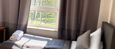A neatly made bed with a checkered bedspread and pillows, a small bedside table with a vase of flowers, and a window with curtains.