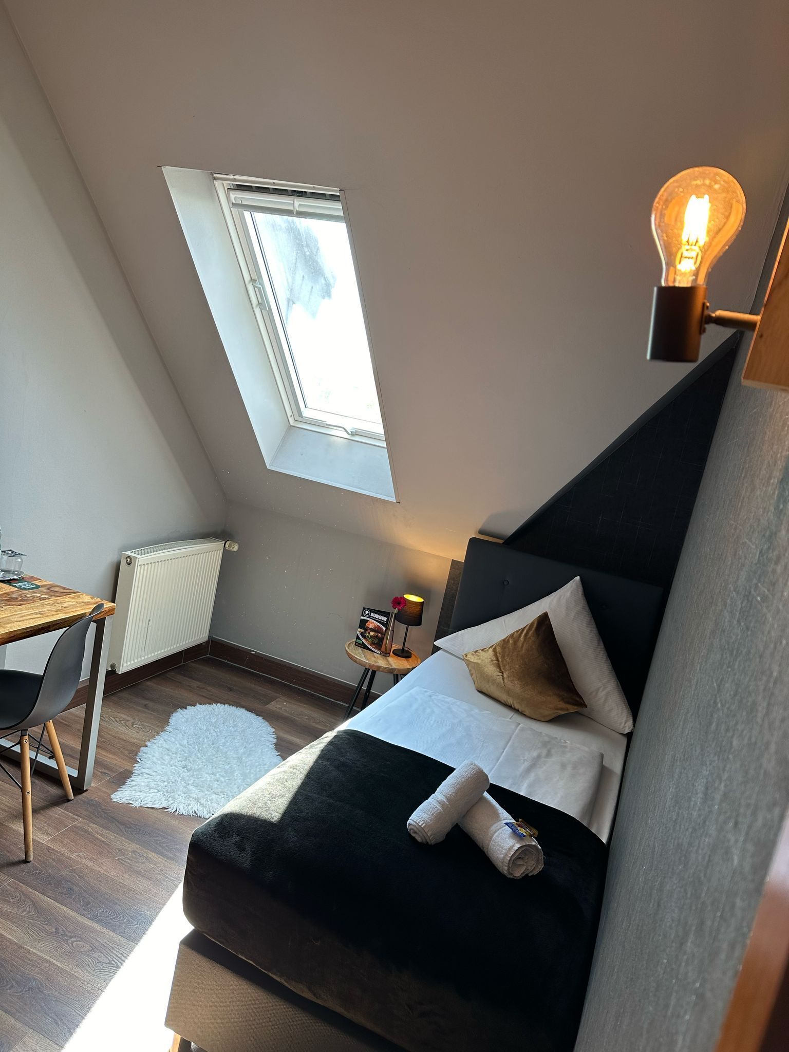 Ein gemütliches Schlafzimmer im Loft-Stil mit schrägem Dach, einem Dachfenster, einem modernen Bett, einem kleinen Tisch mit Lampe und einer Heizung.