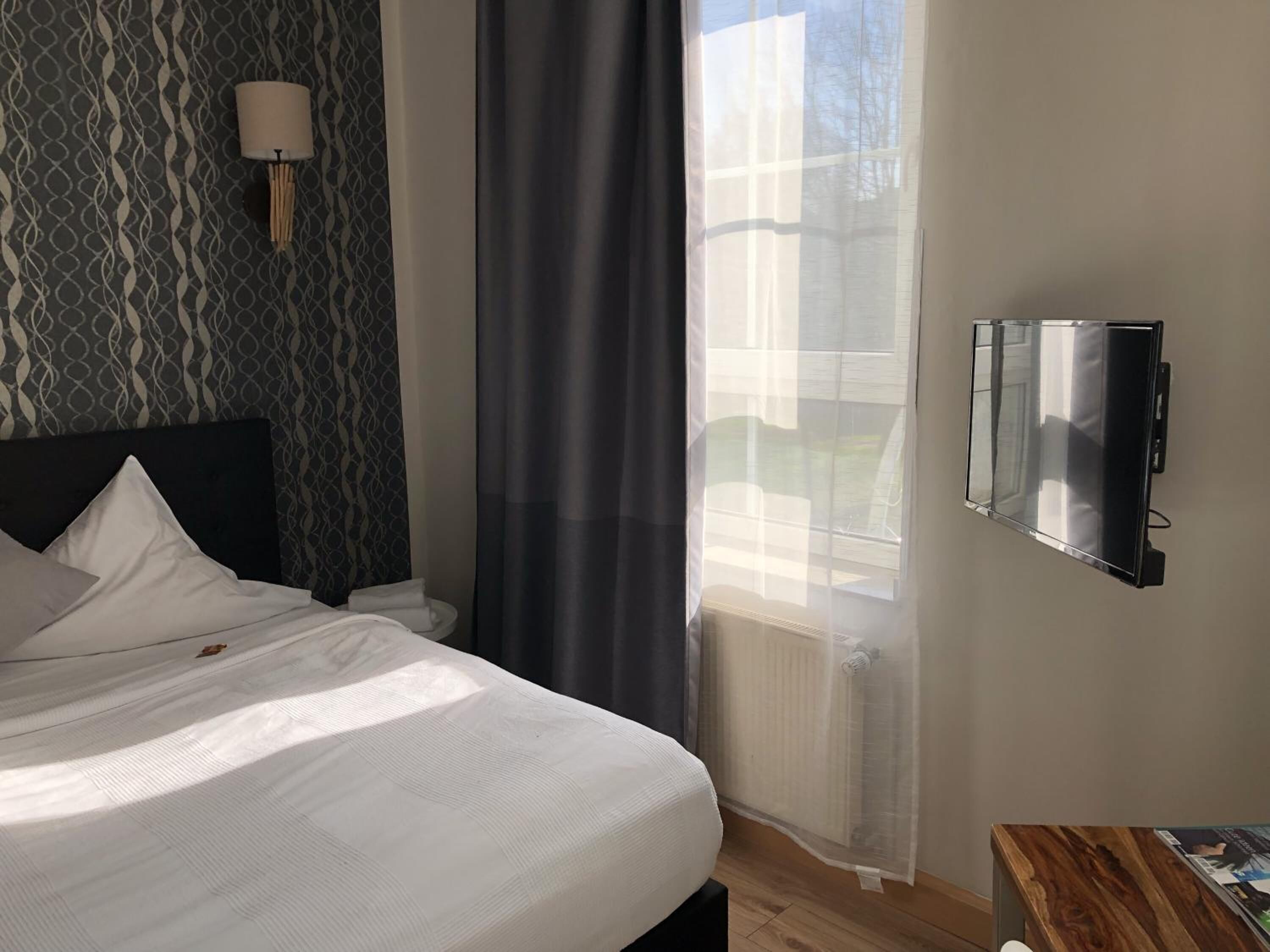Ein Hotelzimmer mit einem Bett, einem an der Wand montierten Fernseher, einem Fenster mit durchsichtigen Vorhängen und einem strukturierten Wandpapier.