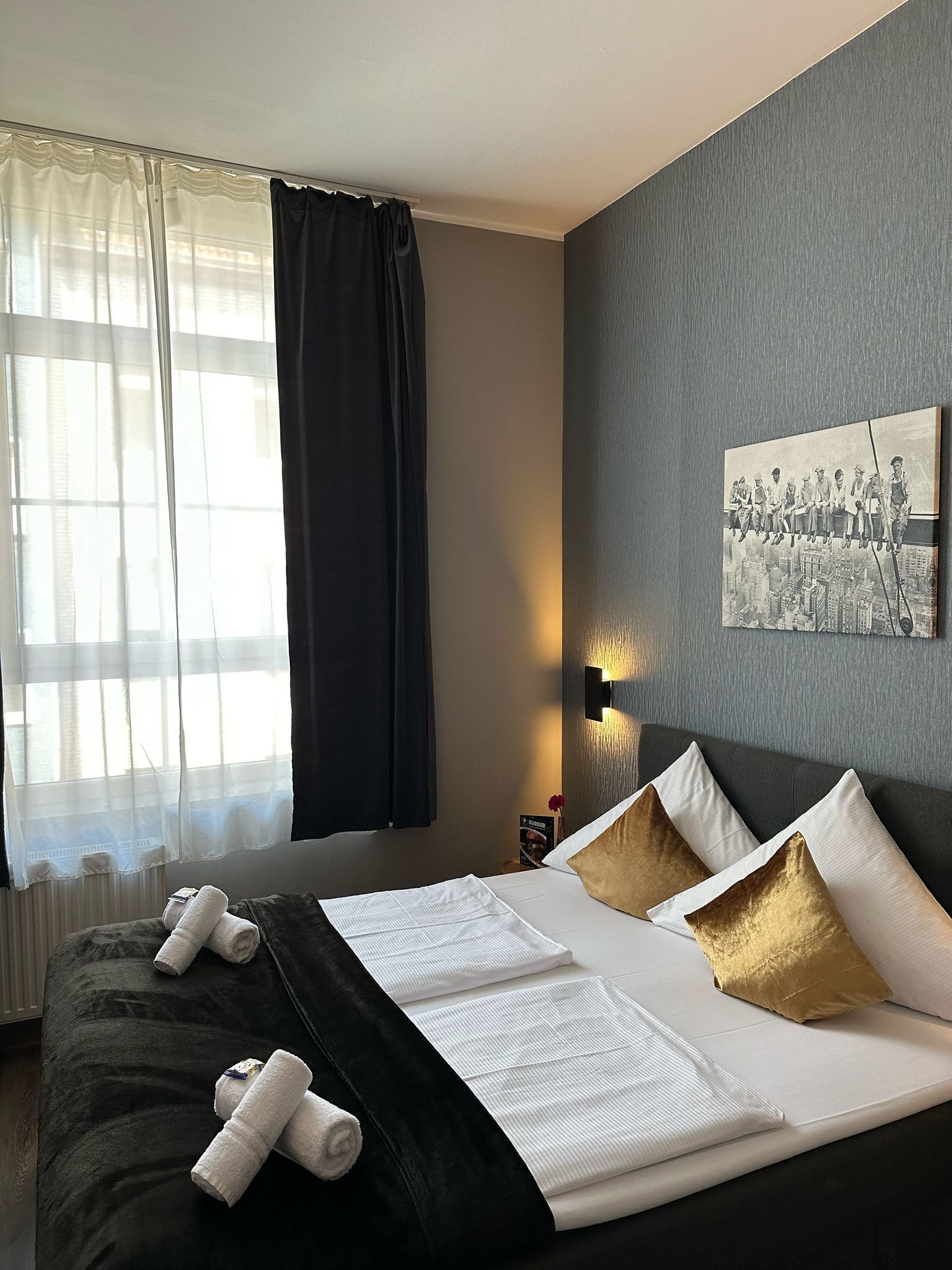 Una habitación de hotel con una cama, un ventanal con cortinas translúcidas, un cuadro colgado en la pared y una mesita de noche con una lámpara.