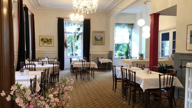 A dining area with multiple tables and chairs, a chandelier, and windows with curtains. - Residence Brunner (Wengen)