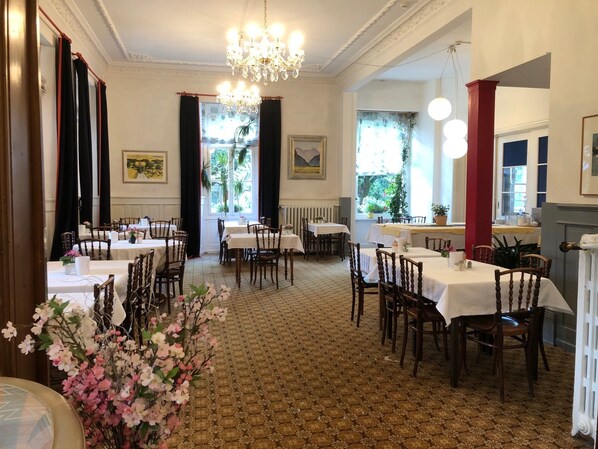 A dining area with multiple tables and chairs, a chandelier, and windows with curtains. - Residence Brunner (Wengen)