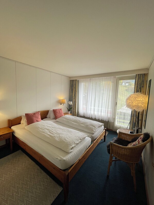 A hotel room with two beds, a nightstand, a chair, and a large window with curtains. - Residence Brunner (Wengen)