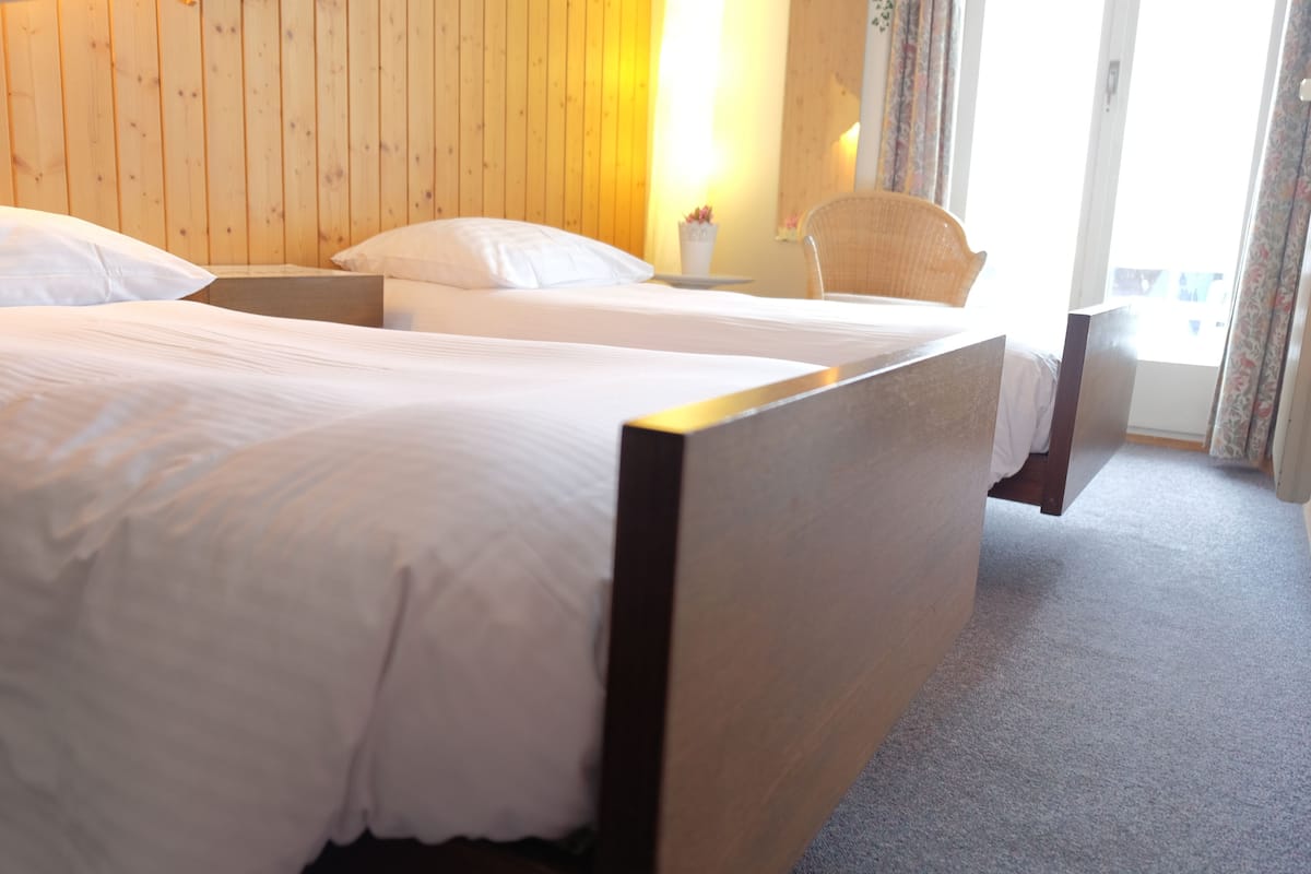 A hotel room with two beds, wooden headboards, a small table with a vase, and a chair.