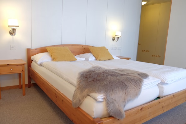 Een houten bed met witte lakens en twee gele kussens, een nachtkastje en twee wandlampen.