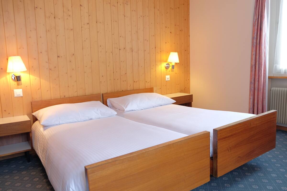 A double bed with wooden headboards and nightstands, two wall-mounted lamps, and a window with curtains.
