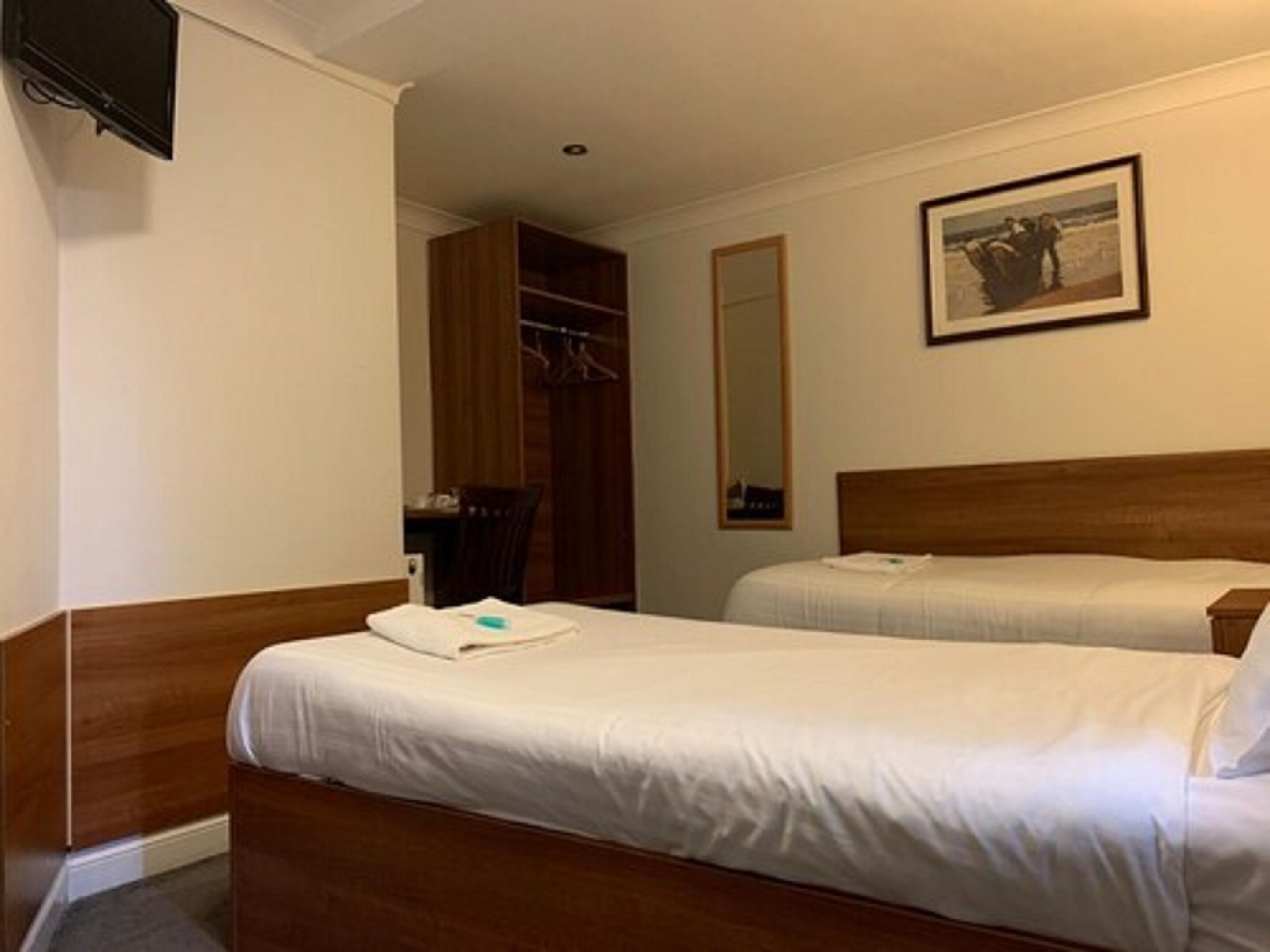A hotel room with two beds, a wooden wardrobe, a television, a mirror, and a framed picture on the wall.