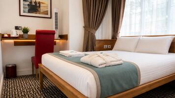 A hotel room with a bed, a desk with a chair, a picture on the wall, and a window with curtains.