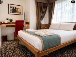 A hotel room with a bed, a desk with a chair, a picture on the wall, and a window with curtains.