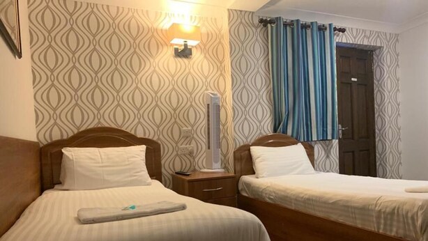 A hotel room with two beds, a bedside table with a lamp, a wall-mounted light, and a door with curtains. - Twickenham Guest House (Twickenham)