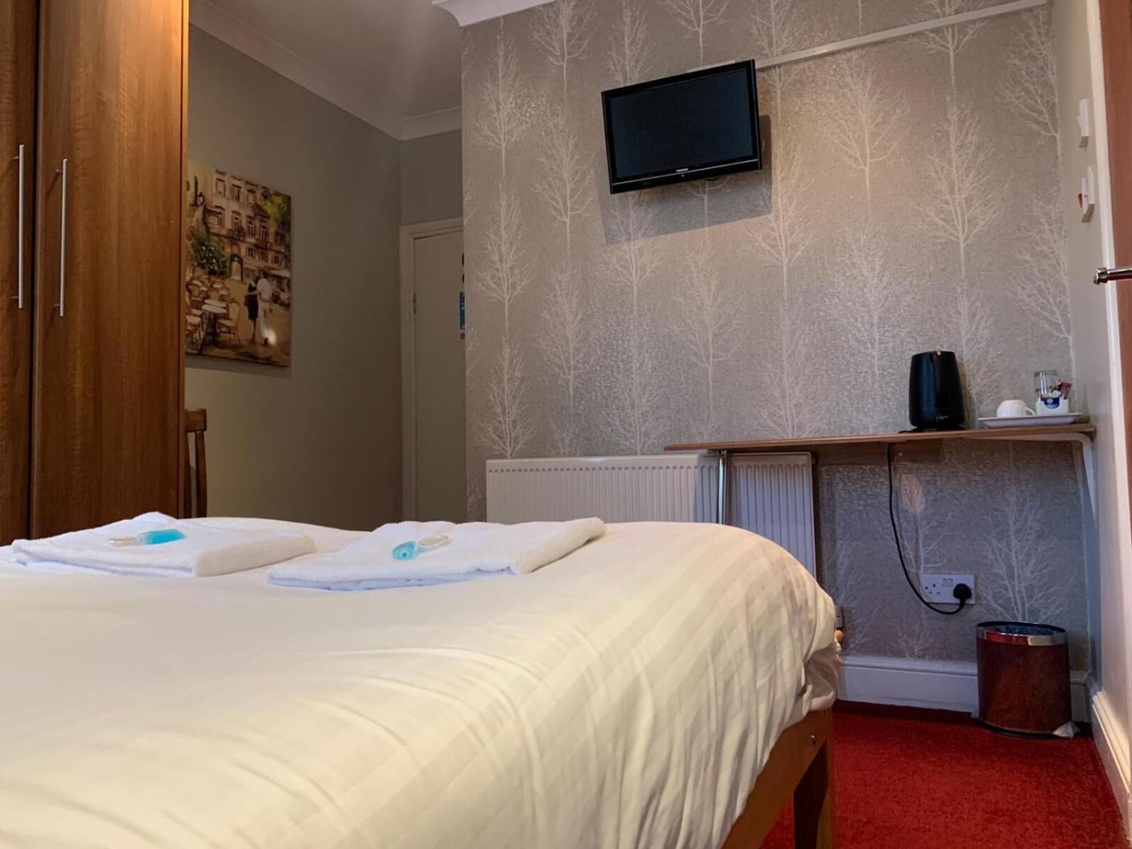 A hotel room with a bed, a television mounted on the wall, a radiator, and a small shelf with a kettle and a cup.