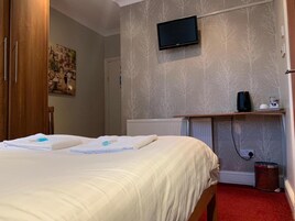 A hotel room with a bed, a television mounted on the wall, a radiator, and a small shelf with a kettle and a cup.