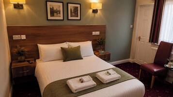 A hotel room with a large bed, two bedside lamps, a headboard, a nightstand with a vase and remote, a chair, and a door with curtains.