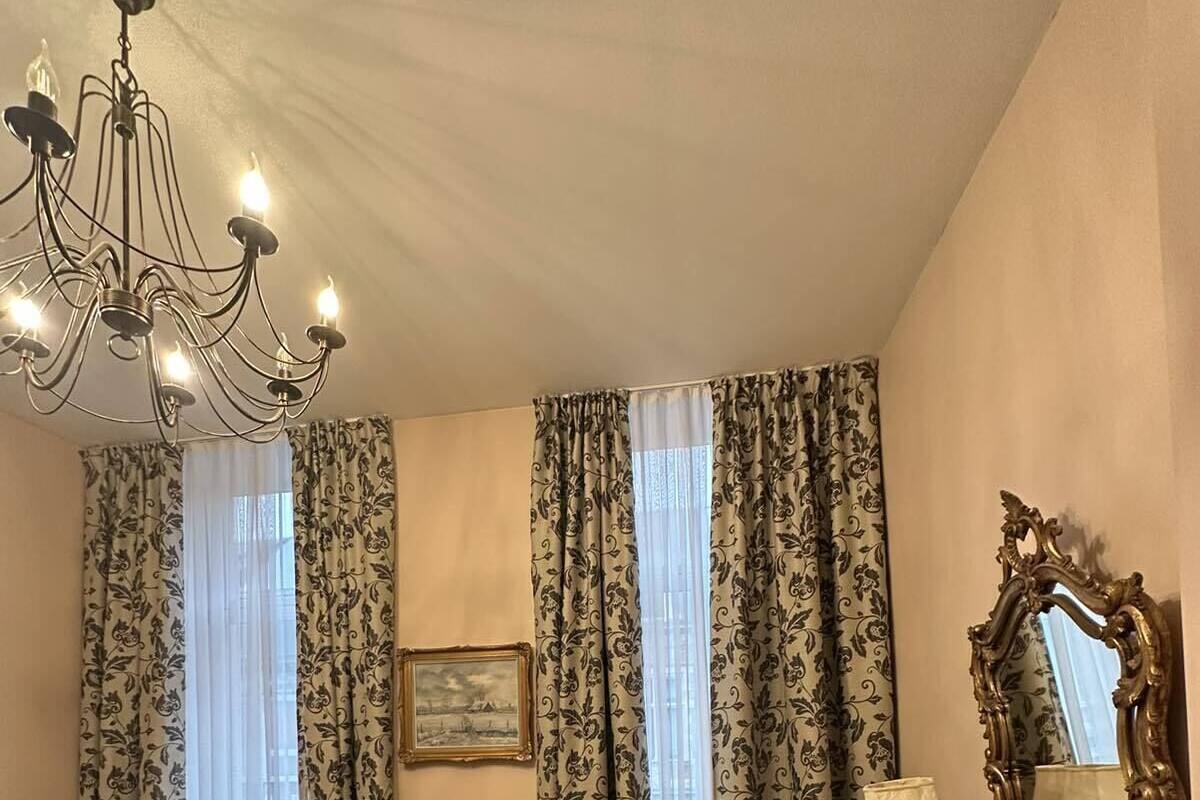 A hotel room with a chandelier, two armchairs, a small table, a desk with a lamp, a large mirror, and a bed with a white cover.