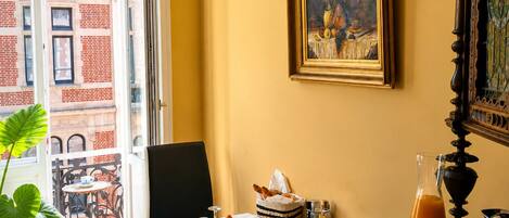 A dining area with a table set for breakfast, a painting on the wall, and a view of a building through an open door.