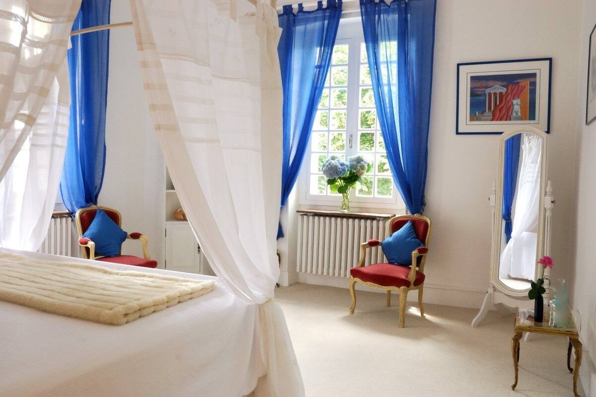 Un dormitorio con una cama, dos sillas, un espejo y una ventana con cortinas azules.