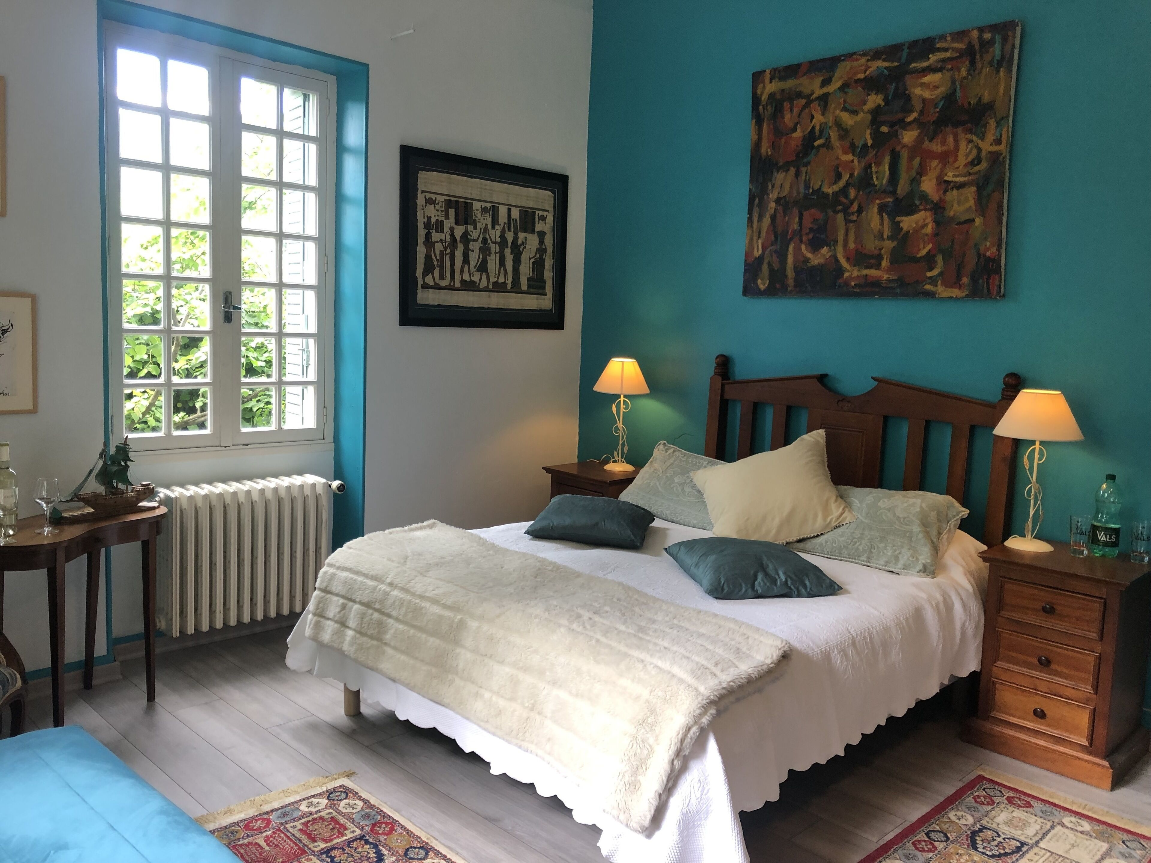 A bedroom with a bed, two bedside tables, a radiator, a window, and a painting on the wall.
