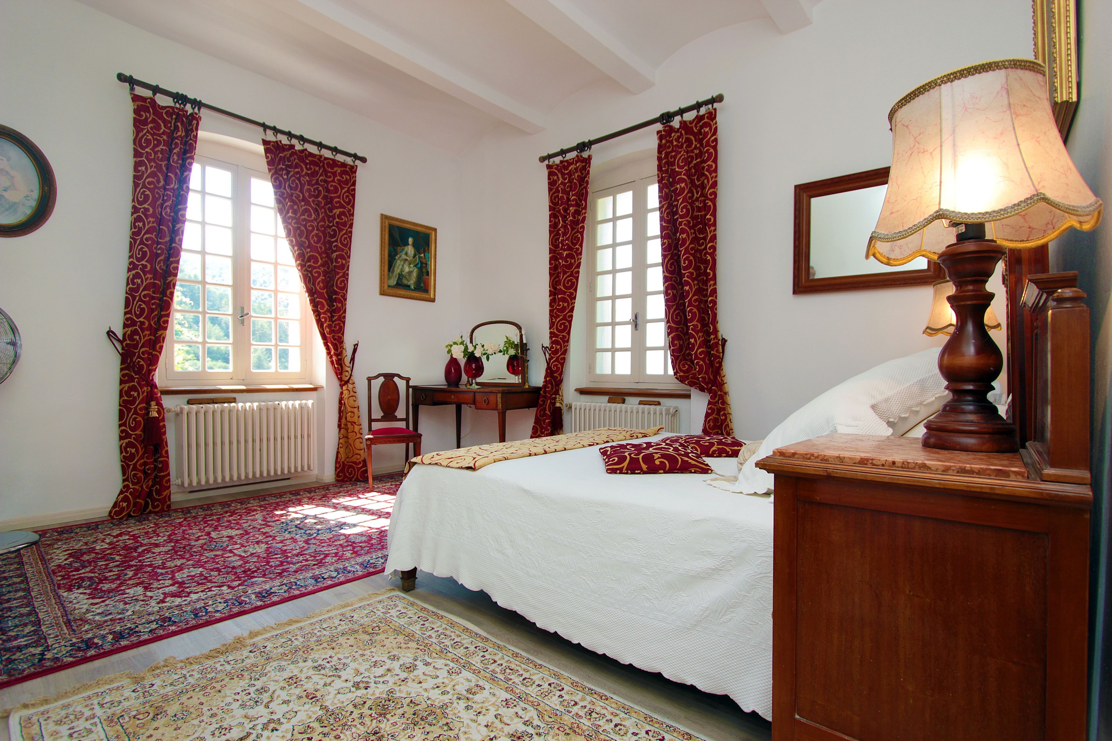 Un dormitorio con cama, mesita de noche, lámpara, ventana con cortinas, radiador, silla, mesa y espejo.