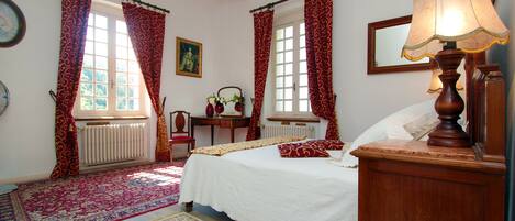 Un dormitorio con cama, mesita de noche, lámpara, ventana con cortinas, radiador, silla, mesa y espejo.