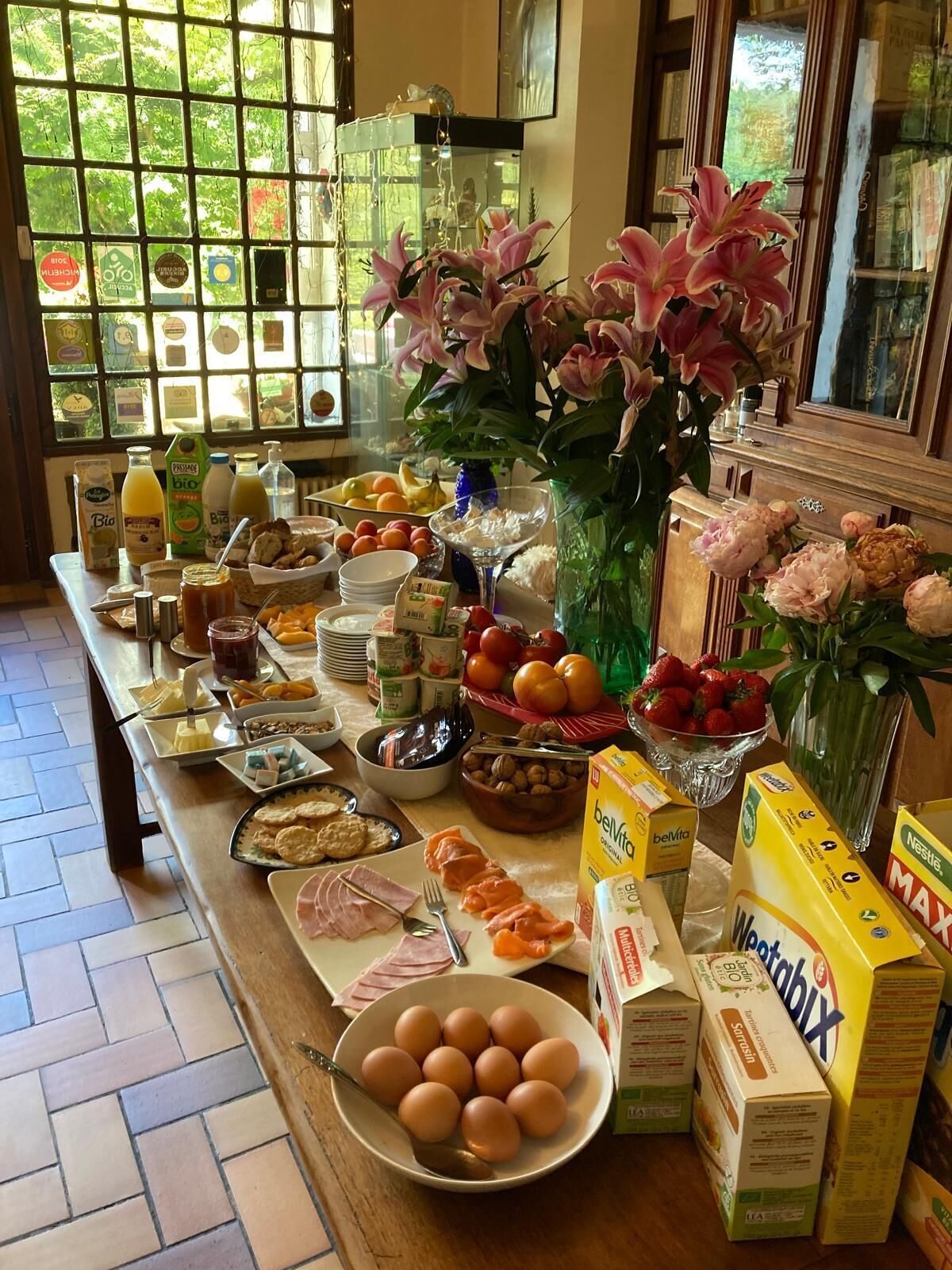 A breakfast buffet with various food items, including eggs, fruits, and beverages, arranged on a wooden table with a view of greenery through a window.