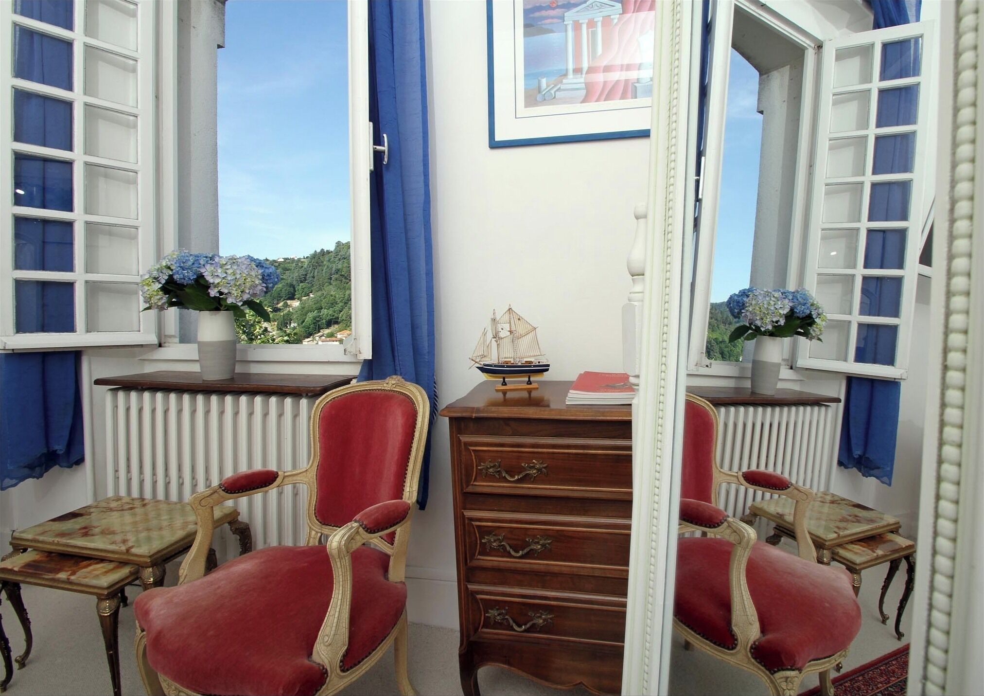 Ein Zimmer mit zwei roten Polstersesseln, einer Holzkommode und einem kleinen Tisch. Zwei Fenster mit blauen Vorhängen und ein gerahmtes Bild an der Wand.