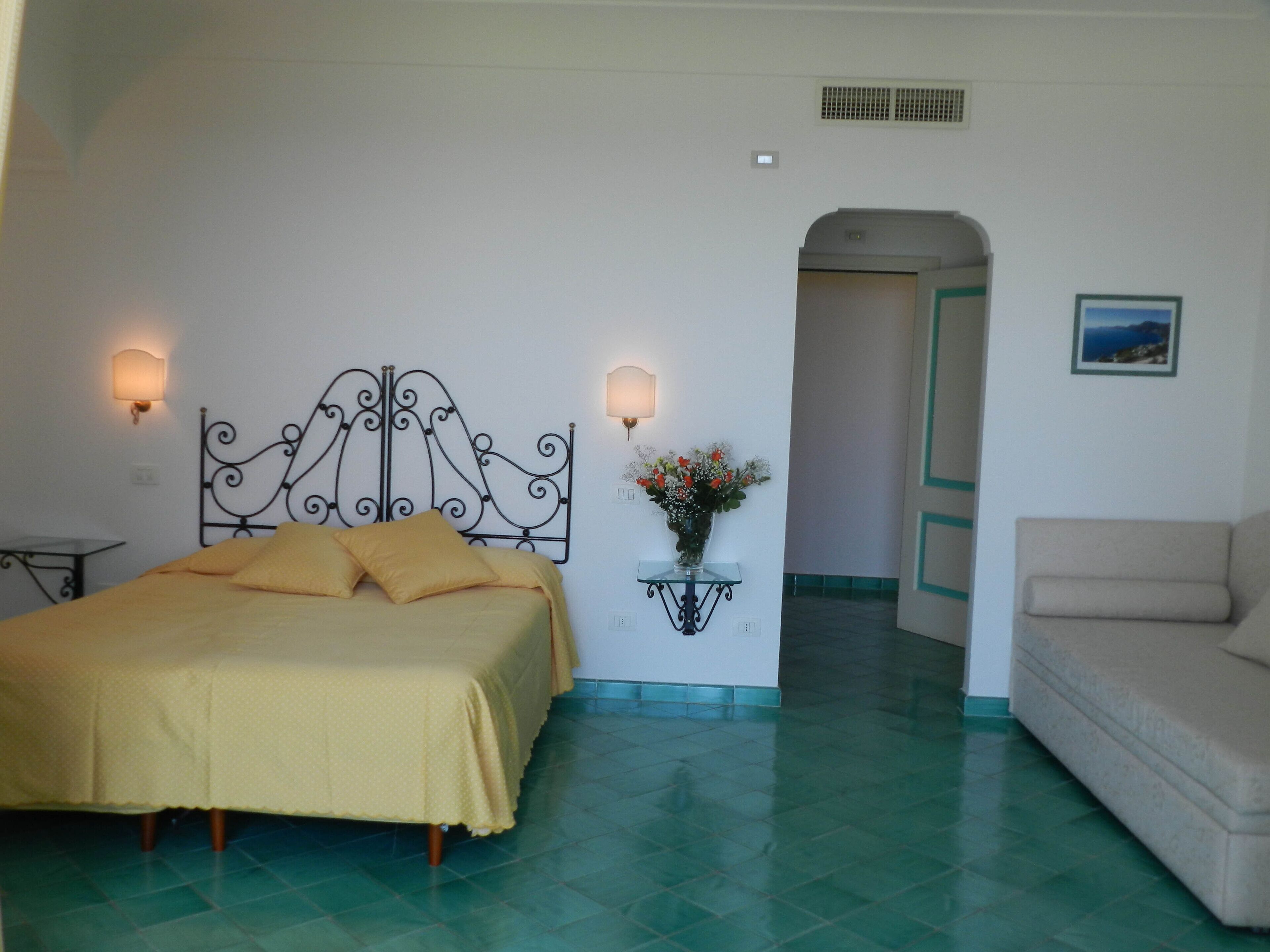 Un dormitorio con una cama, una mesita de noche, una lámpara de pared, un florero con flores y una puerta que da a otra habitación.