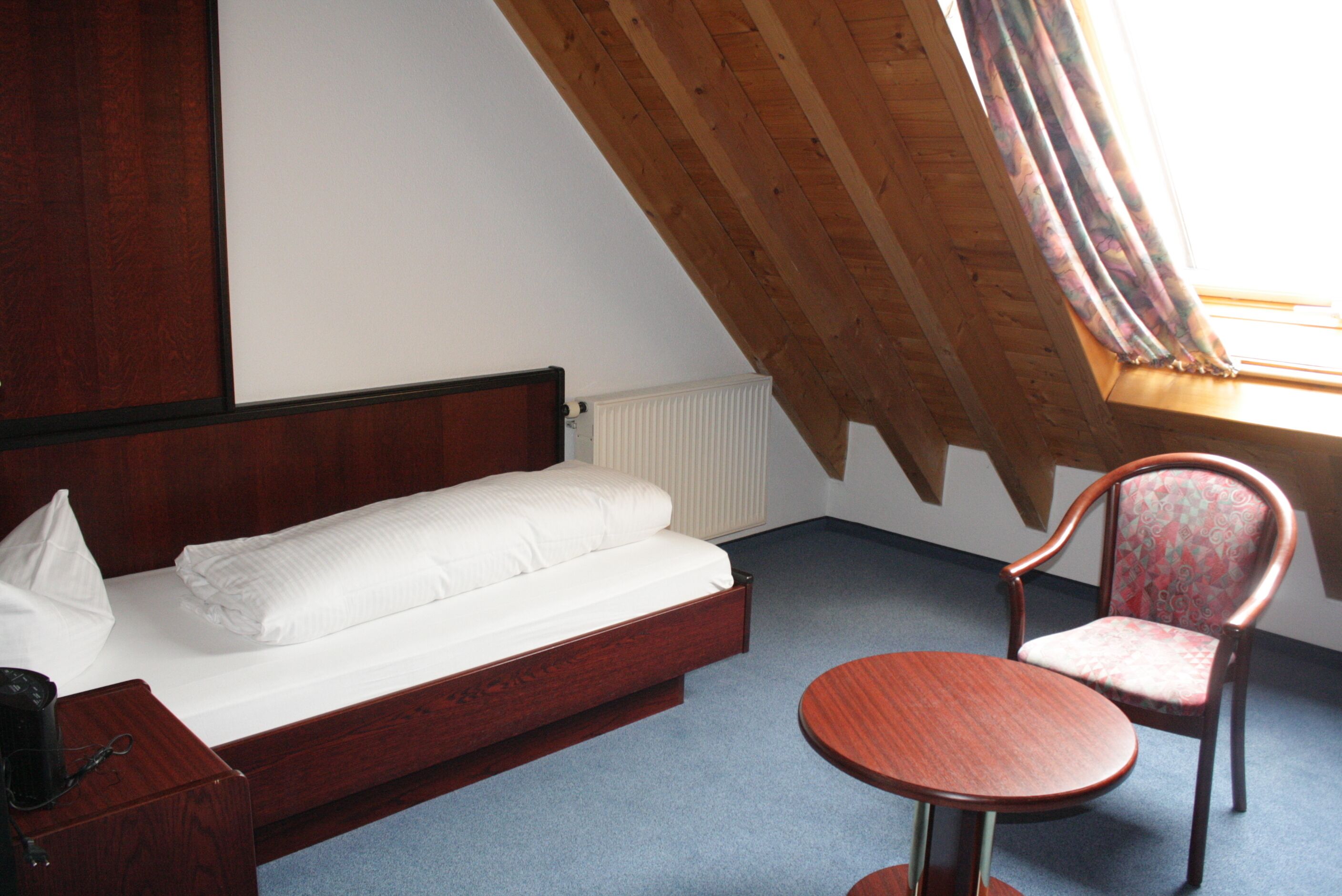 Ein Einzelbett mit Holzgestell, ein kleiner runder Tisch, ein Stuhl und ein Fenster mit Vorhängen.