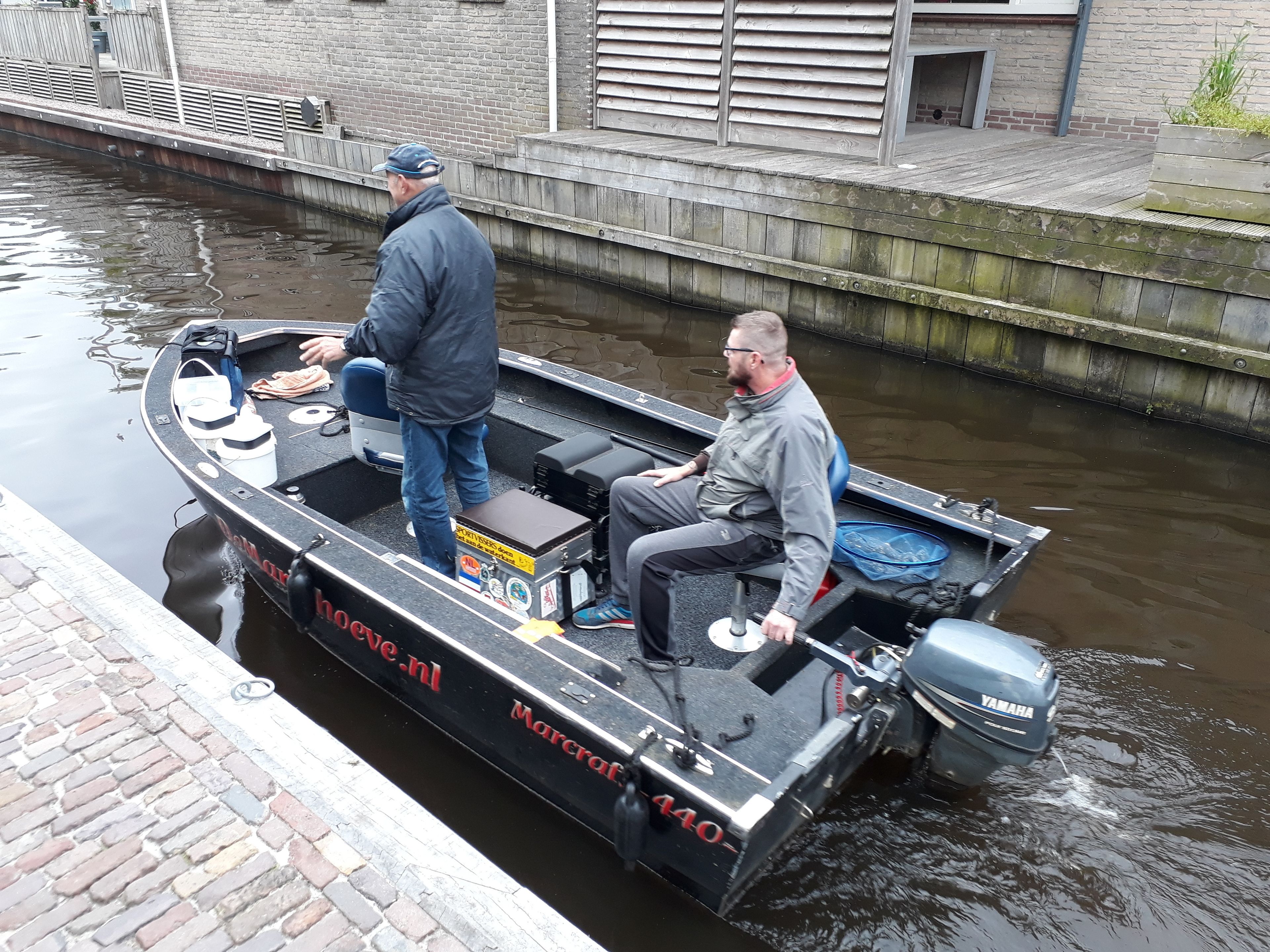 A narrow canal boat with two people, one standing and one seated, equipped with a Yamaha outboard motor.