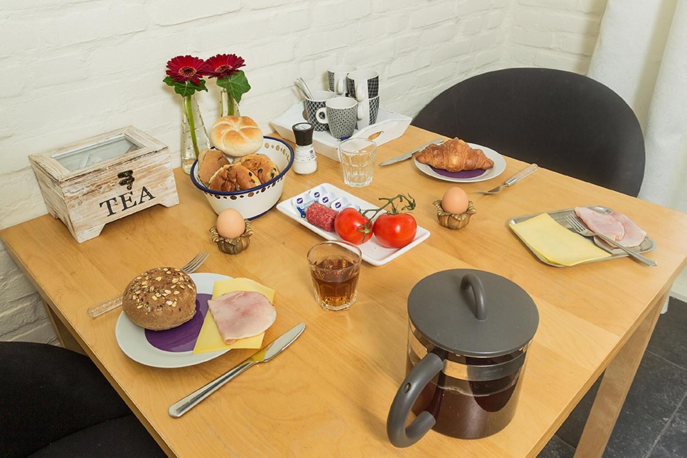 Een ontbijttafel met brood, ham, kaas, tomaten, croissants en een koffiepot.