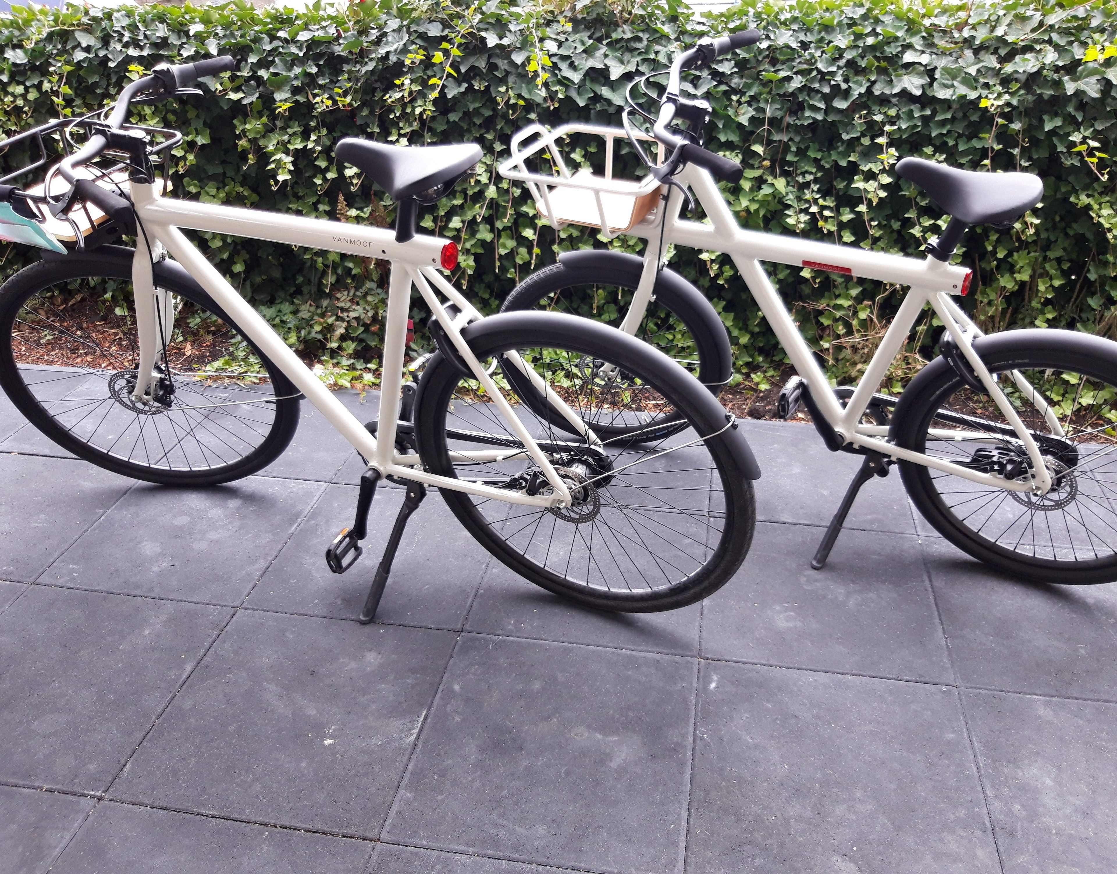 A white bicycle with a basket and a rear rack, parked on a paved surface with a hedge in the background.