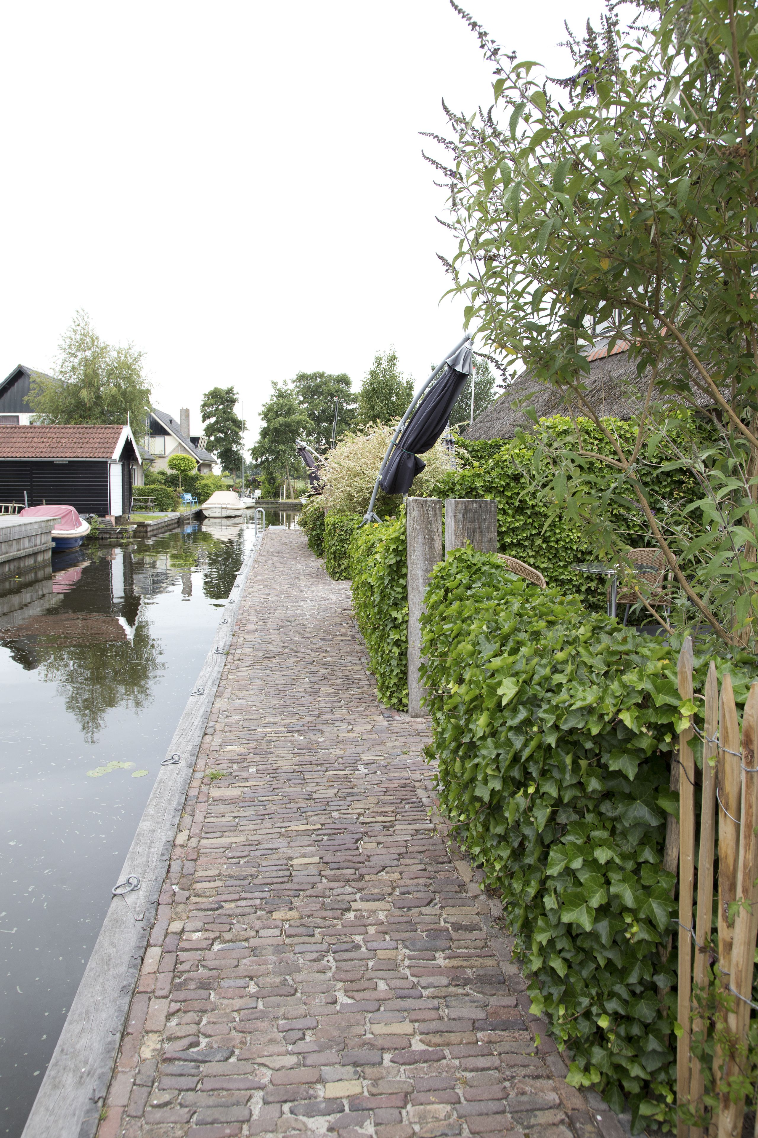 A narrow cobblestone path beside a canal with boats, wooden fences, and greenery.