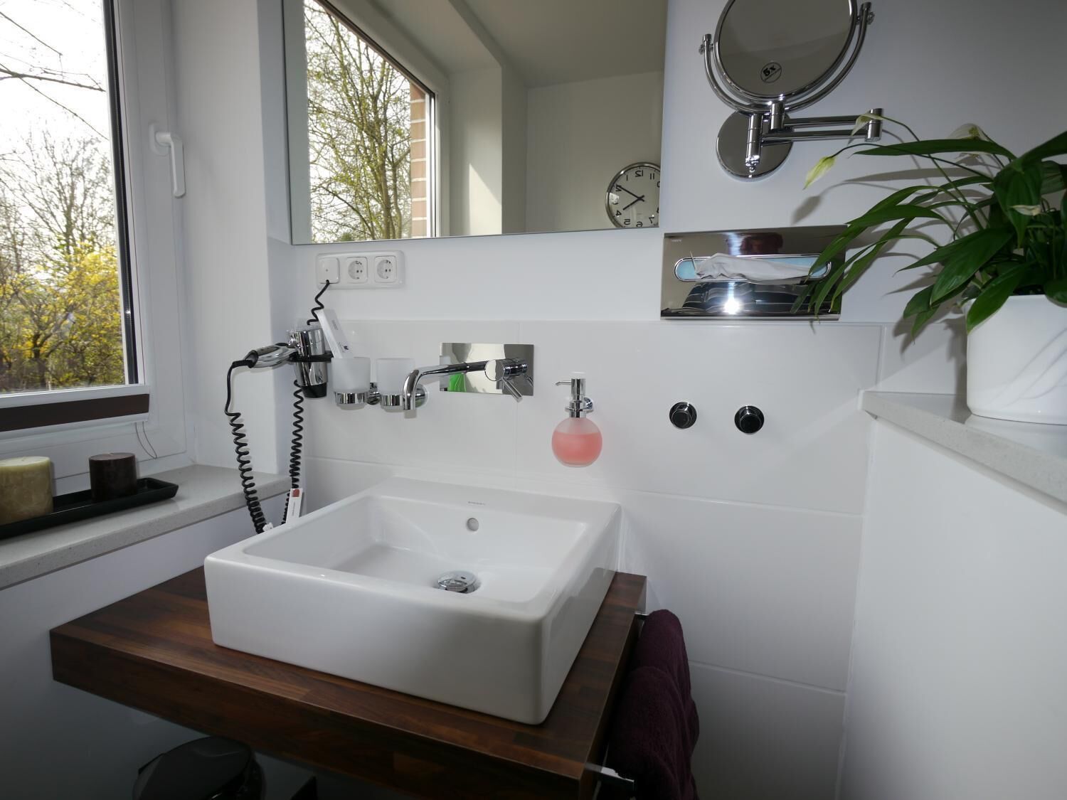 Un baño moderno con un lavamanos blanco, un espejo encima y una ventana con vistas a árboles.