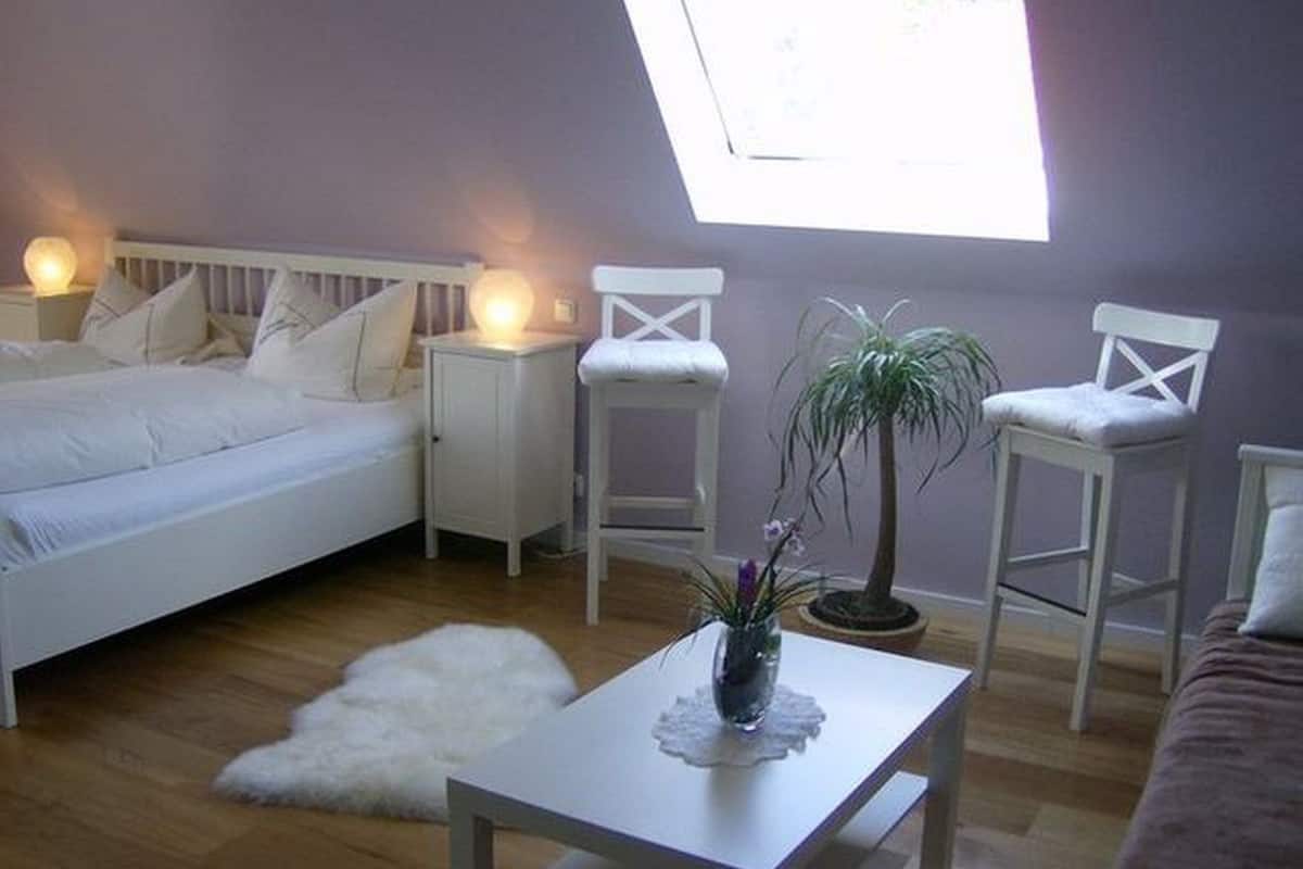 Une chambre à coucher avec un lit, des tables de chevet, une commode, une chaise blanche, une petite table avec une plante et une grande fenêtre.