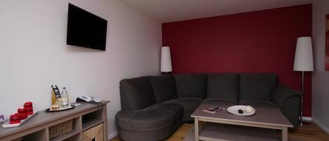 A modern living room with a grey sectional sofa, a small round ottoman, a coffee table, and a flat-screen TV mounted on the wall.