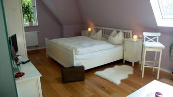 Ein Schlafzimmer mit einem weißen Bett, einem Nachttisch mit Lampe, einem Sessel, einem Schreibtisch mit Computer und einem Fenster mit Pflanze.